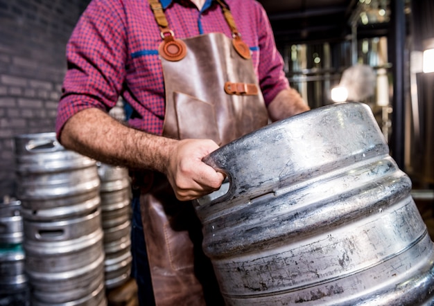 Young male brewer in leather apron holds barrel with craft beer at modern brewery factory