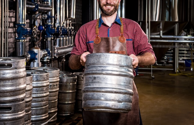 Молодой пивовар в кожаном фартуке держит бочку с крафтовым пивом на современном пивоваренном заводе