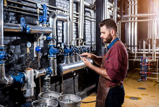 Young male brewer in leather apron supervising the process of beer fermentation at modern brewery fa...