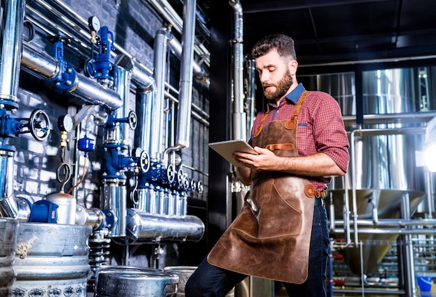 Young male brewer in leather apron supervising the process of beer fermentation at modern brewery fa...