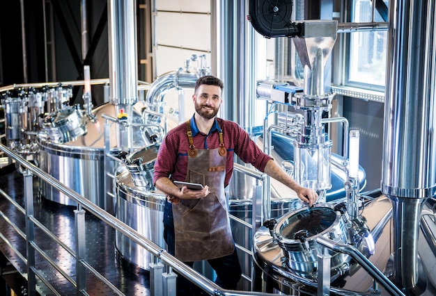 Foto giovane birraio maschio in grembiule in pelle che supervisiona il processo di fermentazione della birra nella moderna fabbrica di birra fa...