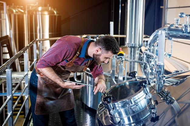 Young male brewer in leather apron supervising the process of beer fermentation at modern brewery fa...