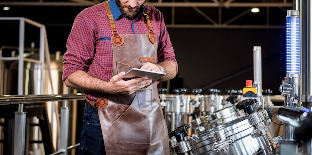 Young male brewer in leather apron supervising the process of beer fermentation at modern brewery fa...