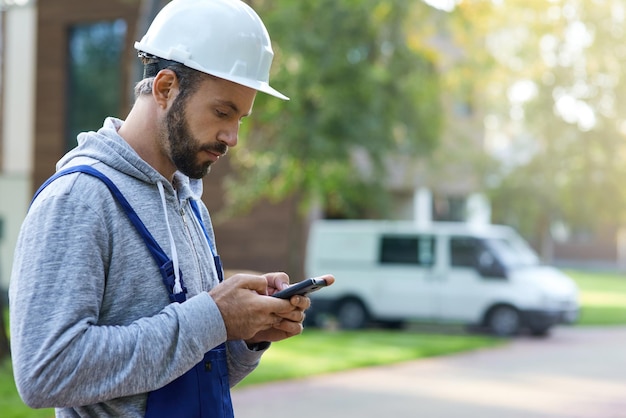 Young male builder in hardhat using smartphone standing outdoors while working at cottage