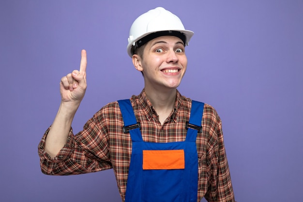 Young male builder wearing uniform isolated on purple wall