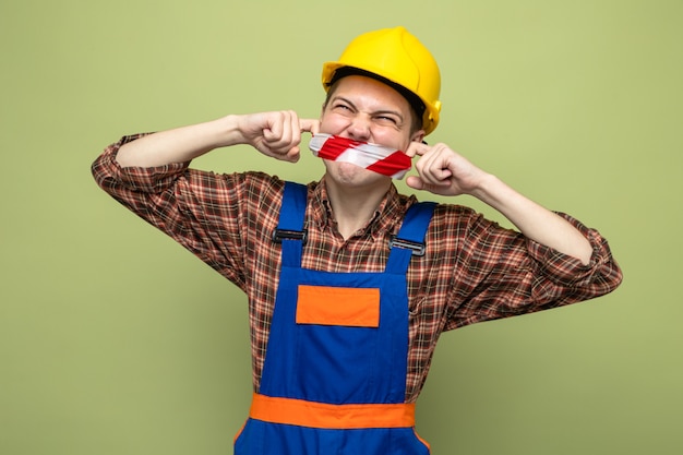 Young male builder wearing uniform sealed mouth with duct tape\
isolated on olive green wall