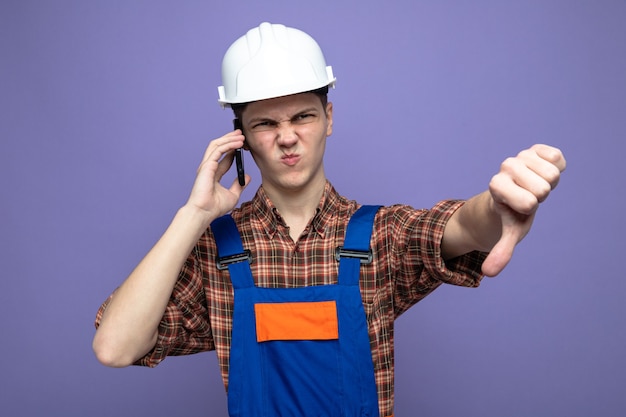 Young male builder wearing uniform speaks on phone isolated on purple wall