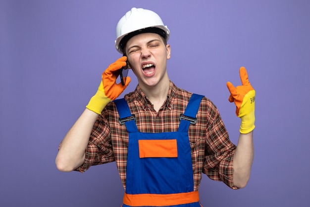 Young male builder wearing uniform with gloves speaks on phone isolated on purple wall