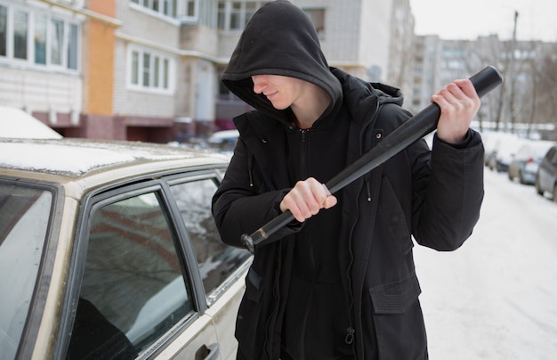 A young male bully in a black jacket with a baseball tries to break a car window.