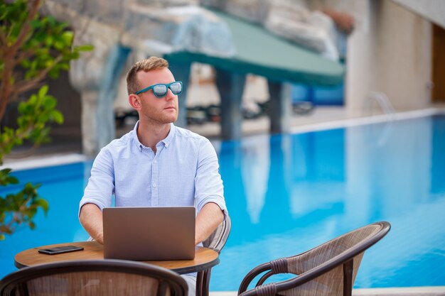 Young male businessman in sunglasses works at a laptop sitting at a table near the pool. Remote work. Freelancer