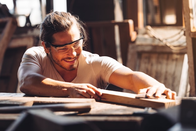 A young male carpenter builder working overall equals a wooden bar with a milling machine in the workshop, 