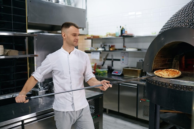 Young male chef holding freshly baked pizza on long shovel. Selective focus of hot Italian fast food