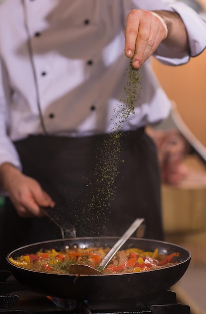 Young male chef putting spices on vegetables in wok at commercial kitchen