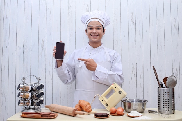 Young male chef in white toque and apron hold cellphone with blank screen for promotional content.