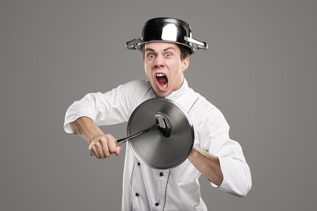 Young male cook with saucepan on head screaming battle cry at camera and hitting lid with ladle against gray background
