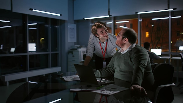 Young male coworkers discuss and work on laptop together late in dark office