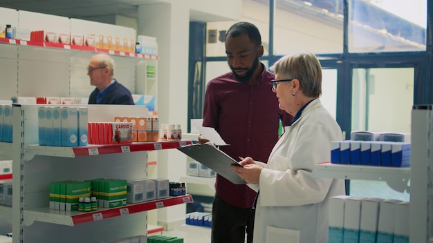 Young male customer giving prescription paper to assistant to find disease treatment and box of medicaments. Person looking at supplements packages on pharmaceutics store shelves.