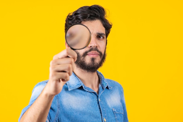 Young male detective holding magnifying glass by right eye while standing in front of camera in isolation