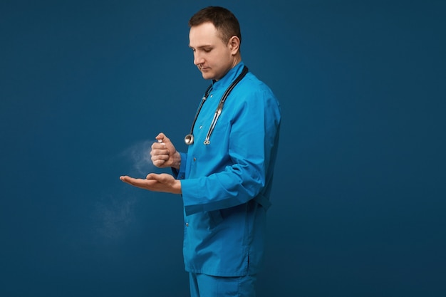 Giovane medico maschio in uniforme medica blu facendo uso dello spruzzo del prodotto disinfettante della mano e posando sul blu
