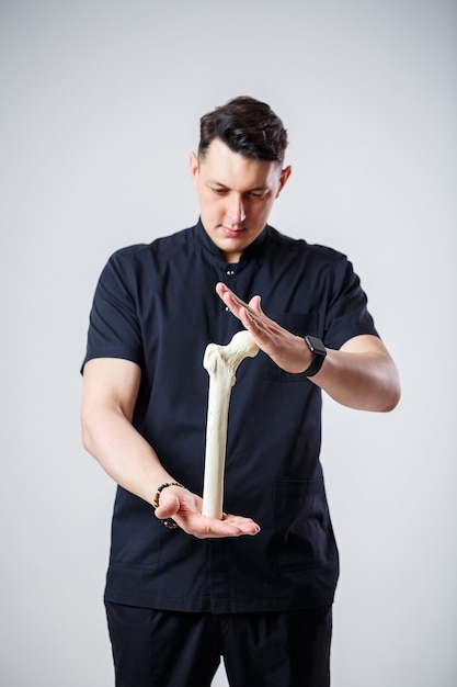 Young male doctor chief physician in a black surgical suit. Close-up portrait. Holds a femur in his hands. Isolated on white background.