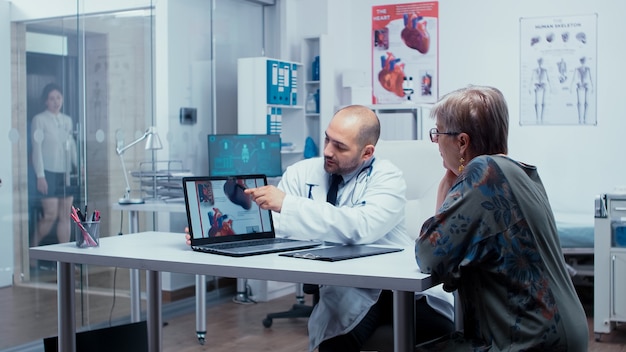 Young male doctor explaining possible heart disease to elderly retired old woman patient. Heart disease problems presented by cardiologist cardiology, heart attach. Health care in modern private clini