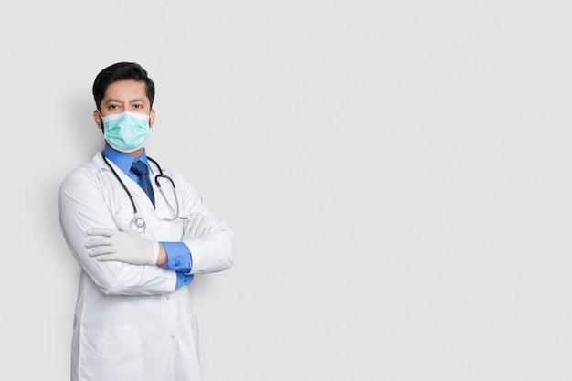 Photo young male doctor face covered with mask and arm cross over isolated wall, health concept