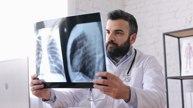 Young male doctor is looking at chest xray while sitting in medical office