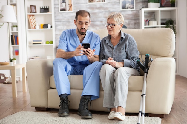 Young male doctor in nursing home helping senior woman to use mobile phone.