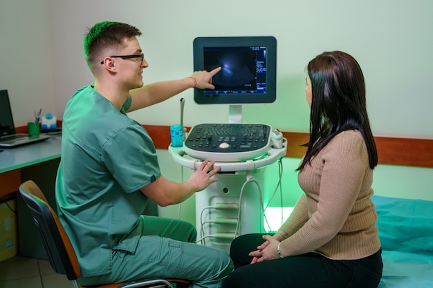 若い男性医師が患者に腹腔の超音波検査を見せます。医者は女性の分析を説明します。特別な設備を備えた元気いっぱいの男性医師。