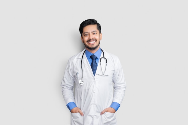 Young male doctor smiling with stethoscope and arm in pockets isolated on white wall. Health insurance concept.