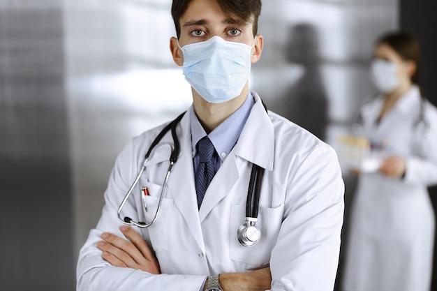 Young male doctor standing with arms crossed in clinic. Smart physician wearing medical protective mask in modern hospital. Medicine and Corona virus concept.