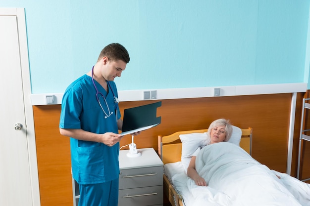 Young male doctor with phonendoscope on his neck looks at the analysis scores of his female patient, who is lying in the hospital bed in the hospital ward. Healthcare concept