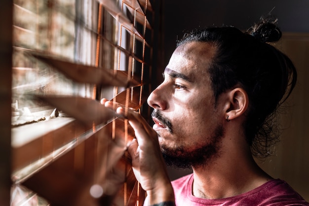 A young male doing self isolation at home to stay safe during Coronavirus outbreak is looking through the window blinds