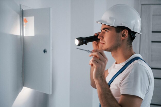 Young male electrician in white hard hat works indoors in the room