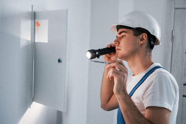 Young male electrician in white hard hat works indoors in the room