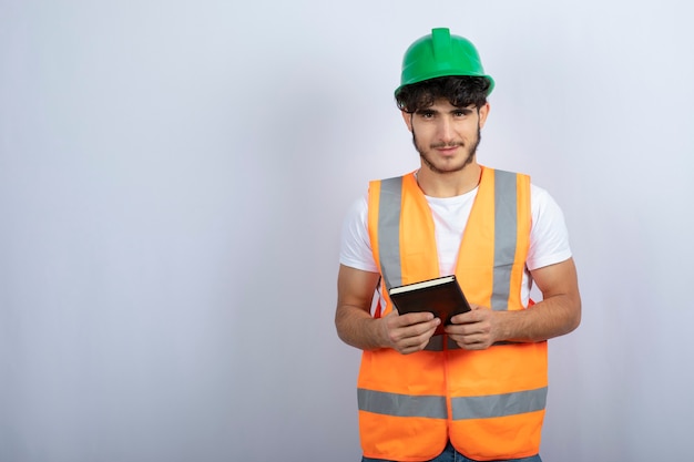 Young male engineer in green hardhat holding notebook on white background. High quality photo