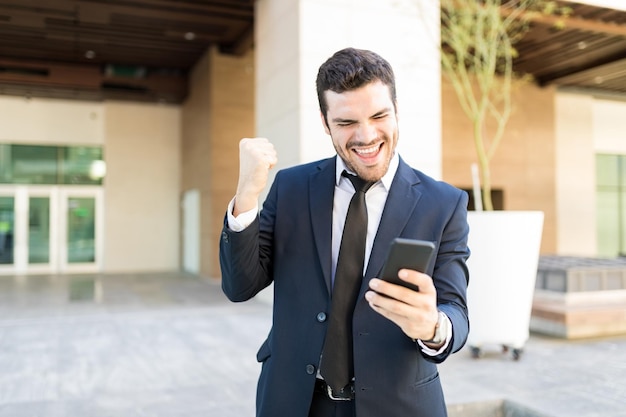 Young male entrepreneur celebrating success while looking at smartphone outside office