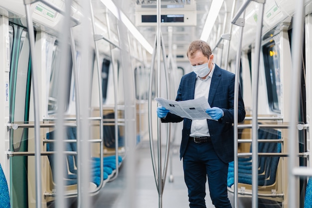 Young male entrepreneur wears disposable medical mask and gloves in metro, prevents coronavirus disease, poses in empty carriage, reads newspaper, finds out information about contagious virus