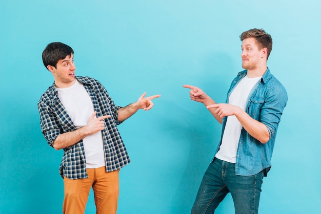 Young male friends making funny faces pointing fingers to each other against blue background