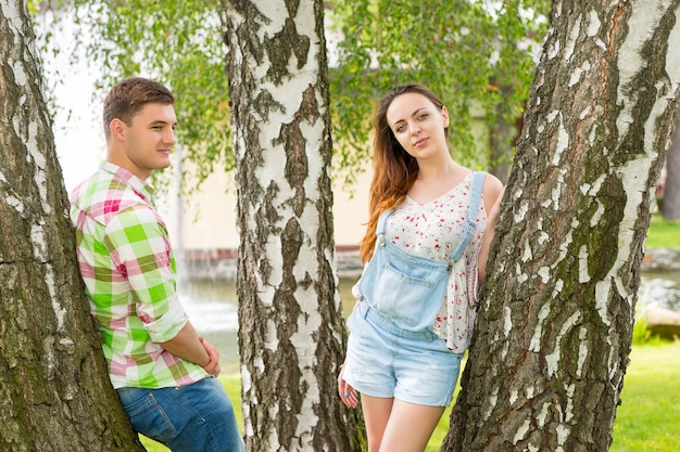 Young male in green and red plaid shirt  and female looking into the camera while leaning on trees in a park with fountain and different trees