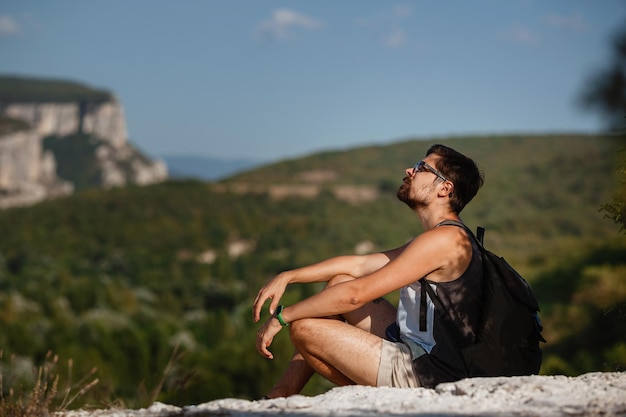 Young male hiker with backpack relaxing on top of a mountain during calm summer sunset