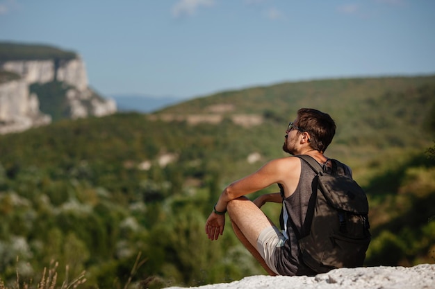 Молодой мужчина-турист с рюкзаком отдыхает на вершине горы во время спокойного летнего заката