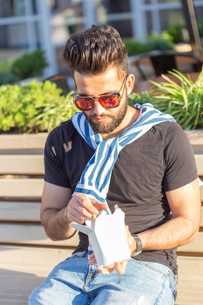 Young male hipster with a mustache and beard is eating chinese noodles with wooden chopsticks from a white lunch box. Asian cuisine concept.