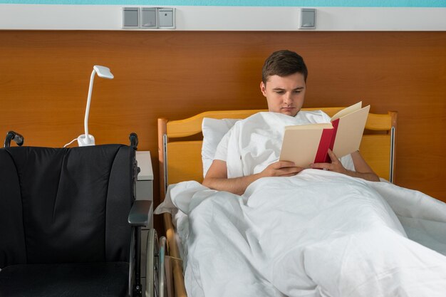 Young male ill patient lying in the hospital bed and reading a book in hospital ward. Healthcare concept