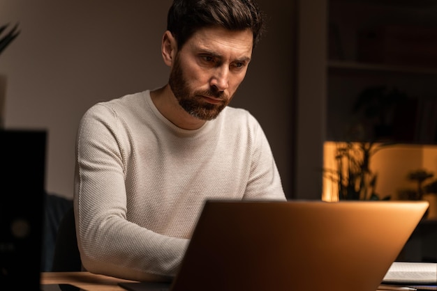 Young male is using laptop in evening at home and looking at digital screen with tired emotions. Stock photo