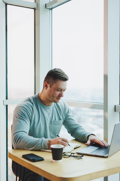 Молодой менеджер-мужчина сидит в офисе с большими окнами и работает за ноутбуком Красивый бизнесмен работает над домашним заданием в кафе