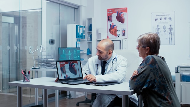 Young male medic presenting a heart booklet on PC to elderly retired senior woman patient. Heart disease problems presented by cardiologist cardiology, heart attach. Health care in modern private clin