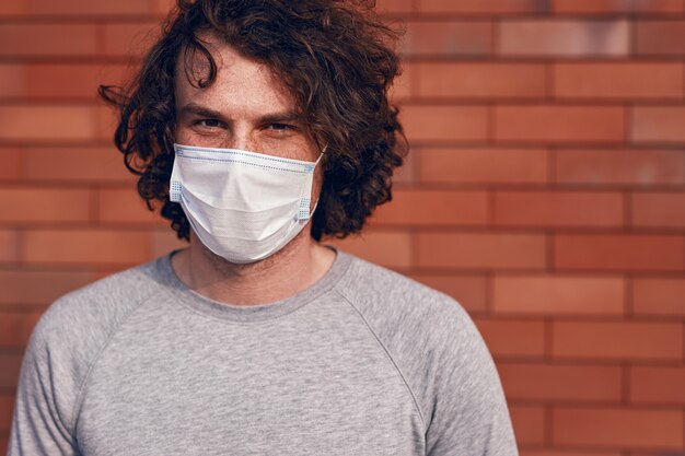 Young male in medical mask looking at camera while standing on blurred background of brick wall during coronavirus epidemic