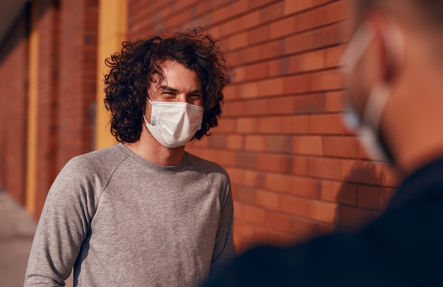 Photo young male in medical mask smiling and communicating with friend while standing near brick wall on city street and keeping social distance during coronavirus epidemic