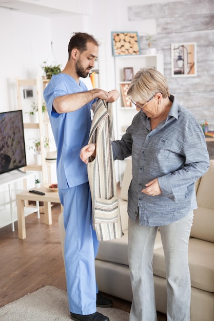 Young male nurse helping senior woman to get dressed in nursing home.